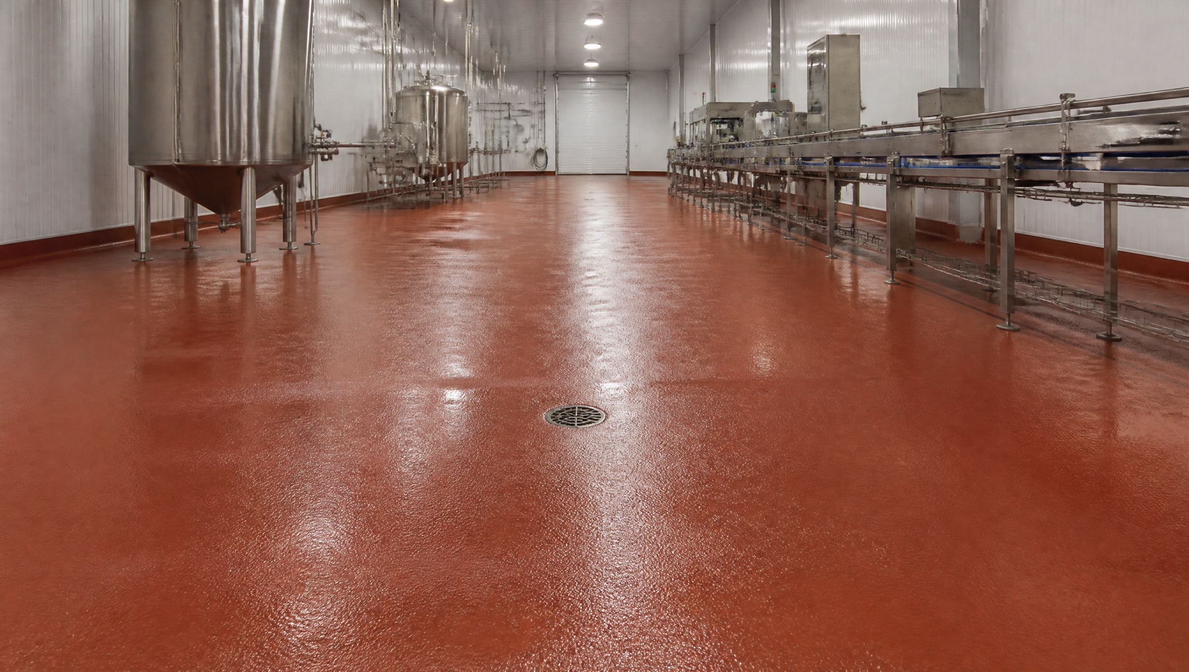 A food processing hall on a red-oxide polyurethane floor, damp from washdown, between steel tanks and a conveyor line